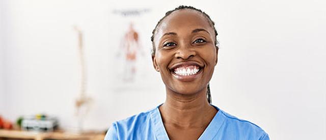 Smiling student dressed in blue scrubs with anatomy poster on wall in background